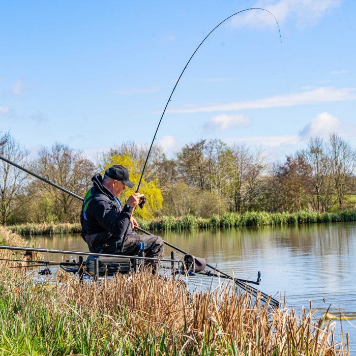 Angler seated on a box playing a fish on a bent feeder rod beside a lake